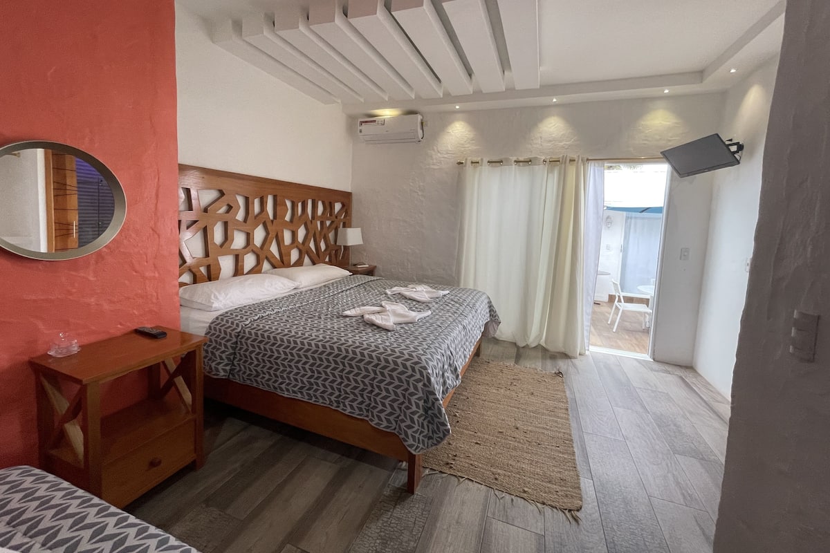 Un dormitorio con cabecera de madera, una mesita de noche con lámpara, cama con colcha estampada y vistas al exterior a través de una ventana con cortinas.