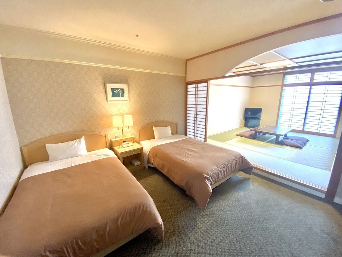 A hotel room with two beds, a nightstand, a lamp, a picture on the wall, and a view through a sliding door.