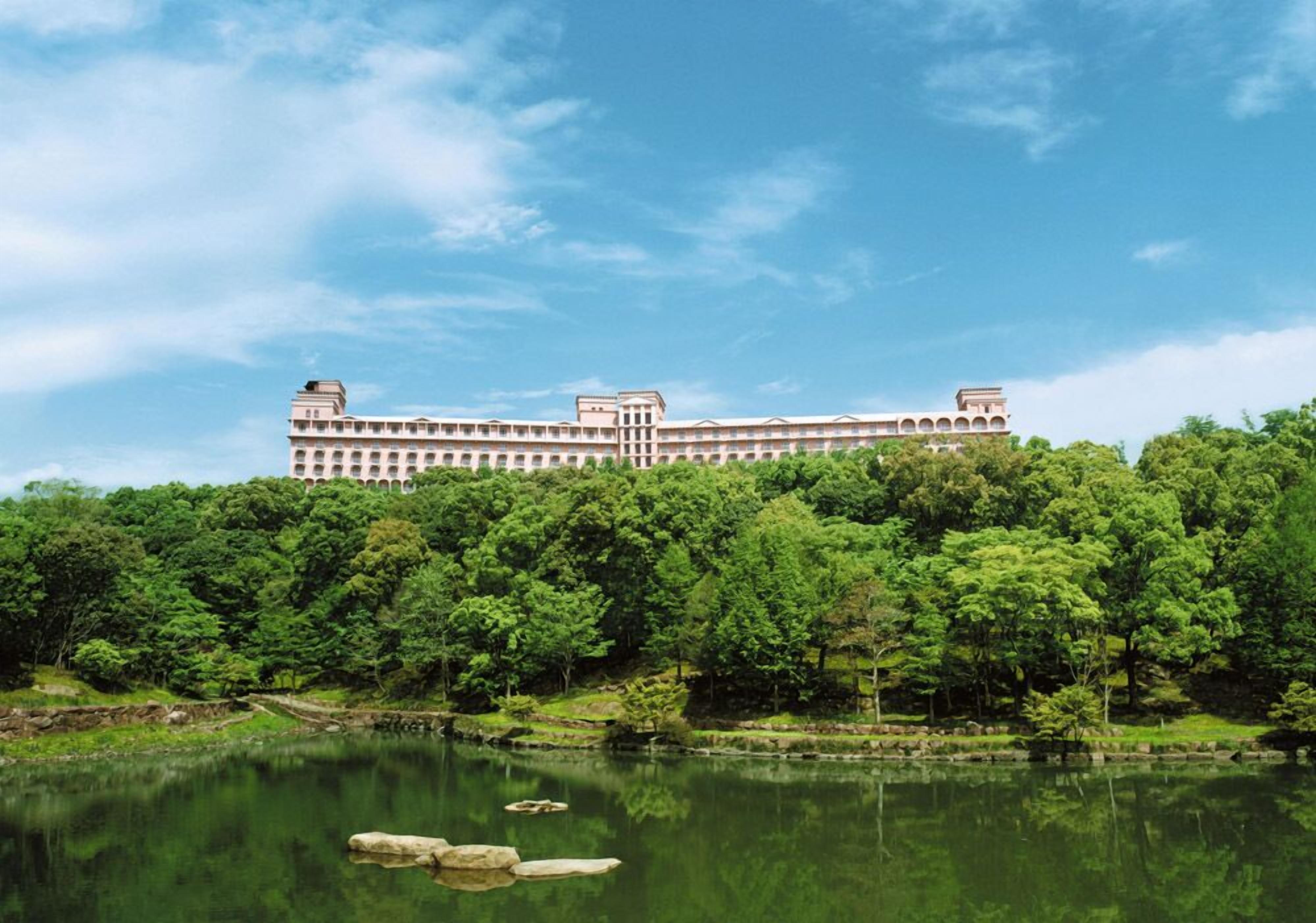 A large, multi-story building with a symmetrical facade, surrounded by lush greenery and a calm body of water.