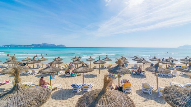 A beach with thatched umbrellas and sun loungers, a clear blue sky, and calm sea.