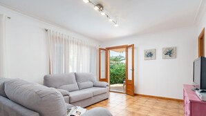 A living room with a grey sofa, a pink cabinet, and a television. There is a door leading to an outdoor area with plants.