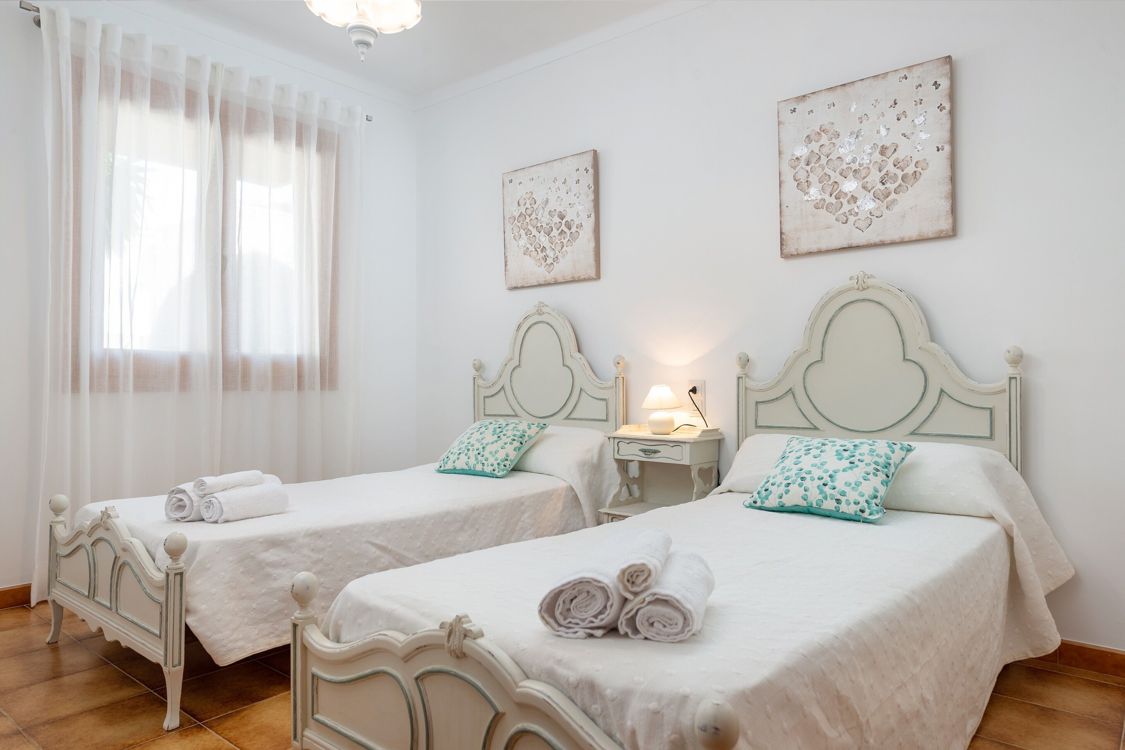 Two twin beds with white linens and rolled towels, a bedside table with a lamp, and a window with sheer curtains.