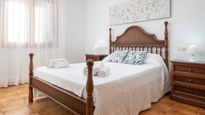A neatly made bed with white linens and rolled towels, a wooden headboard, a bedside table with a lamp, and a wall-mounted abstract artwork.
