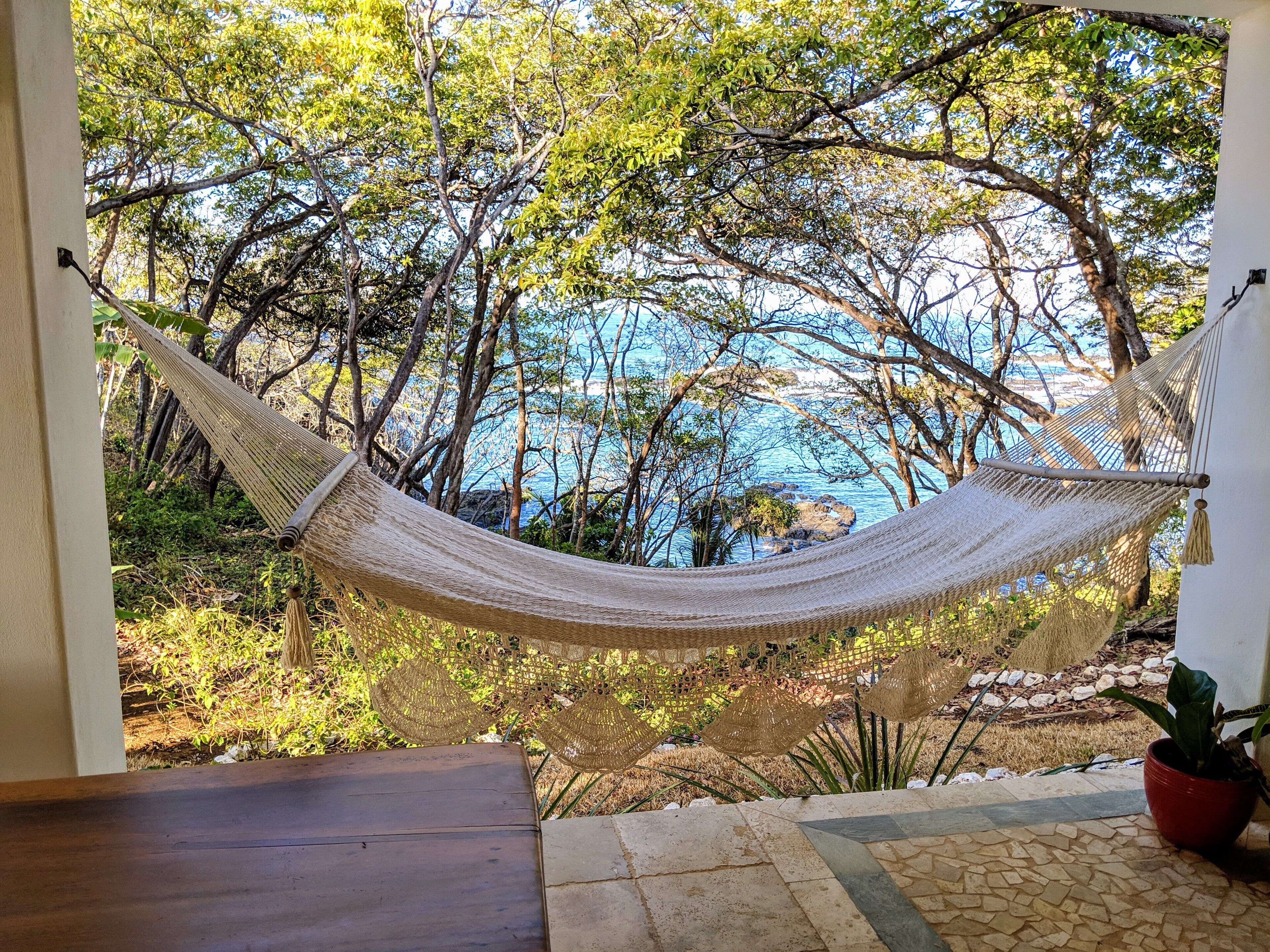 A hammock in a covered outdoor area with a view of the sea and trees.