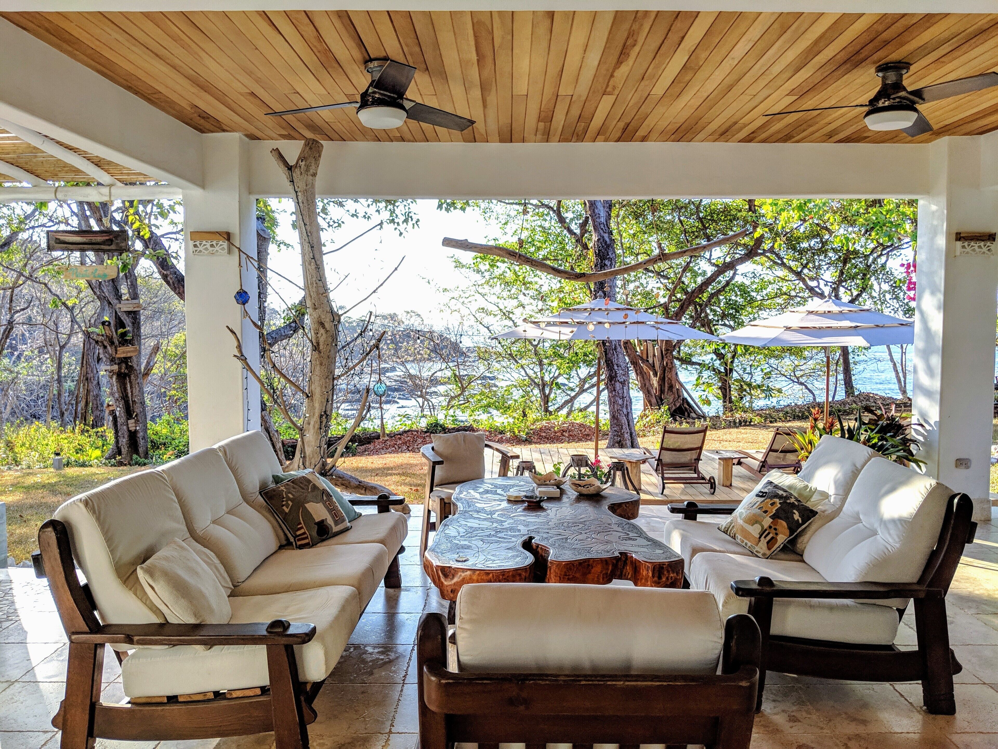 A covered outdoor seating area with a wooden ceiling, white cushioned sofas, and a central coffee table.