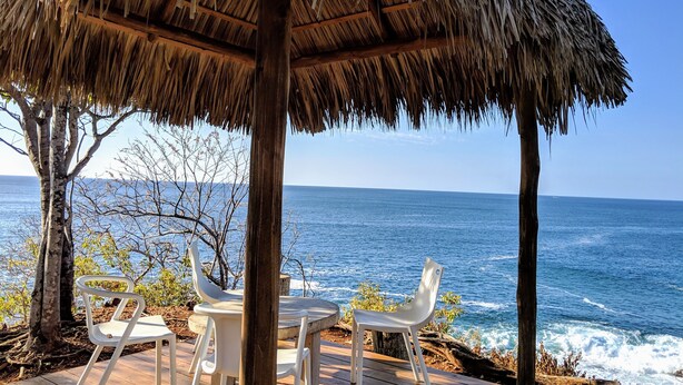 A thatched-roof outdoor seating area with white plastic chairs and a table, overlooking the ocean.