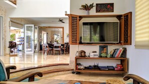 A spacious living area with a wooden dining table, chairs, a television mounted on a wooden cabinet, and a bookshelf with various items.
