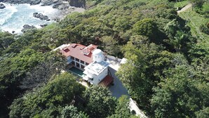 A house with a red roof, surrounded by dense greenery and a rocky coastline.