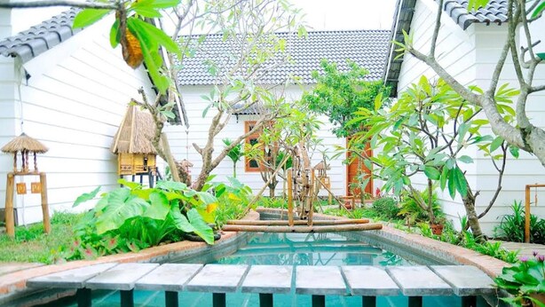 Un área de piscina con una estructura de techo de paja, rodeada de exuberante vegetación y un edificio blanco con techo de tejas.