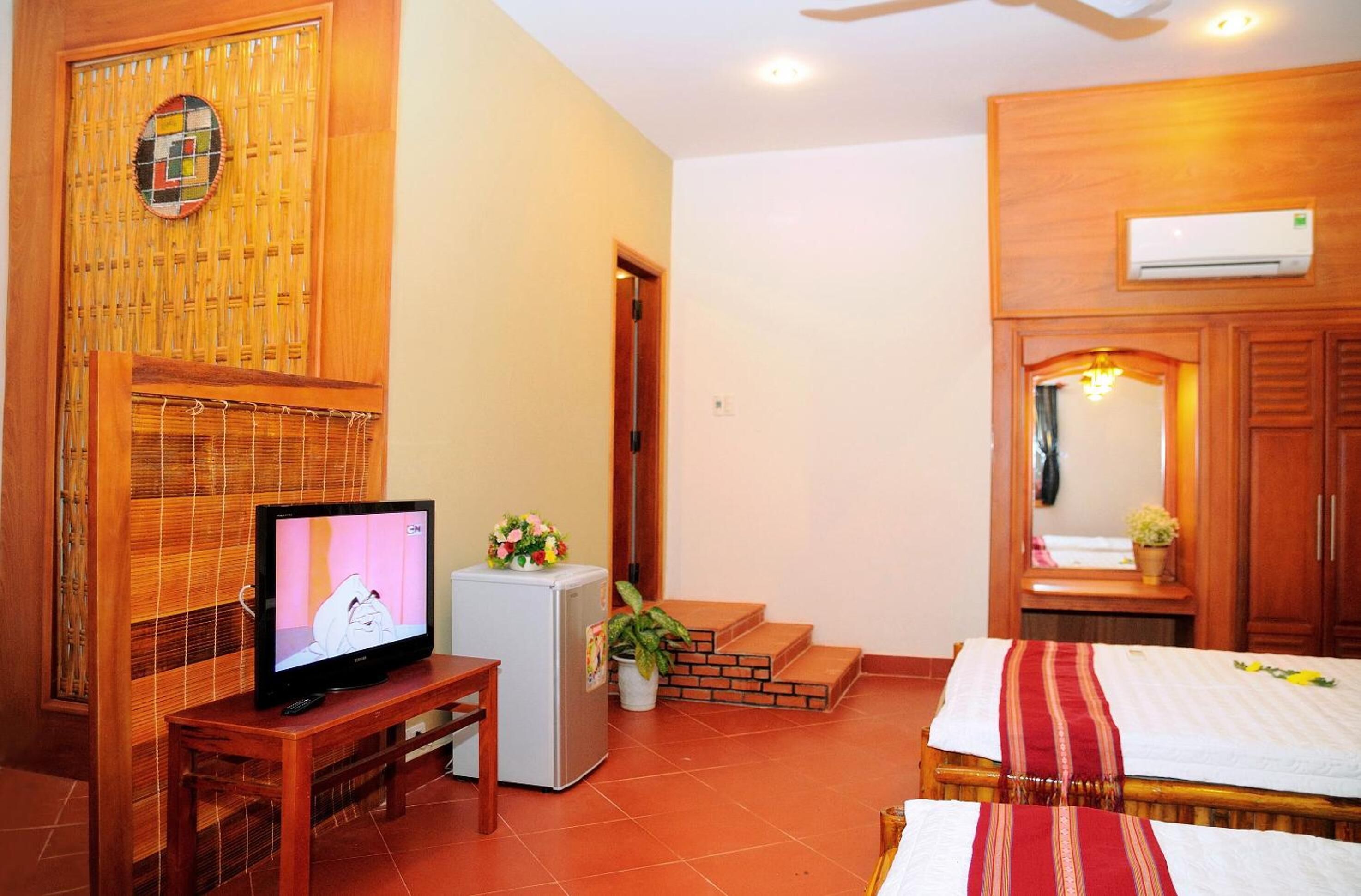 Habitación con televisión sobre un soporte de madera, un refrigerador pequeño, una cama con colcha a rayas rojas y blancas, un armario de madera y un aire acondicionado instalado en la pared.