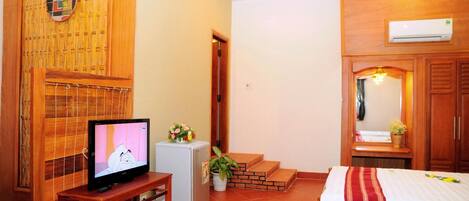 Habitación con televisión sobre un soporte de madera, un refrigerador pequeño, una cama con colcha a rayas rojas y blancas, un armario de madera y un aire acondicionado instalado en la pared.