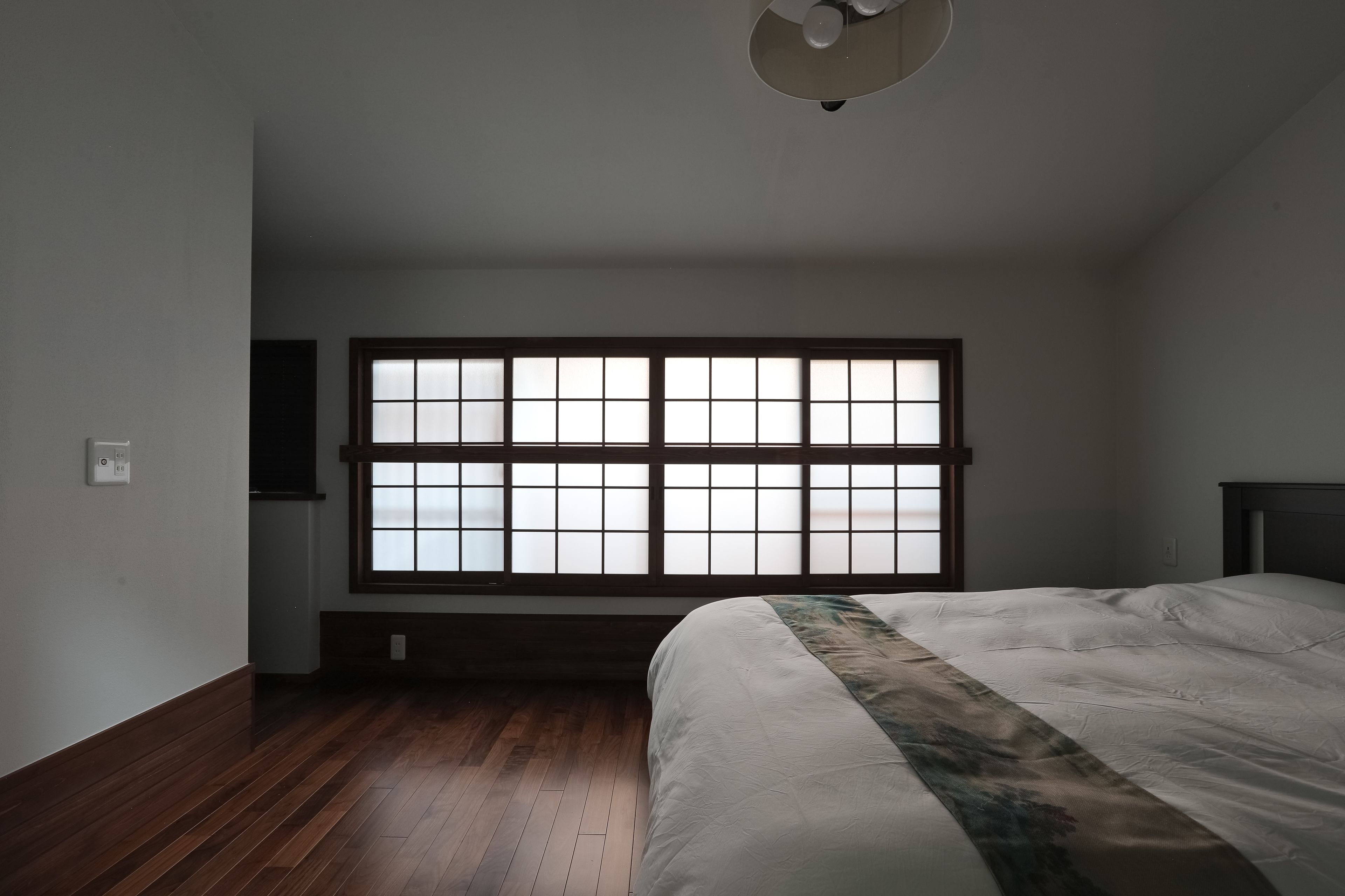 A bedroom with a large window, wooden flooring, and a bed with a patterned bedspread.