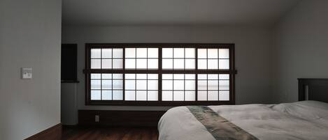 A bedroom with a large window, wooden flooring, and a bed with a patterned bedspread.