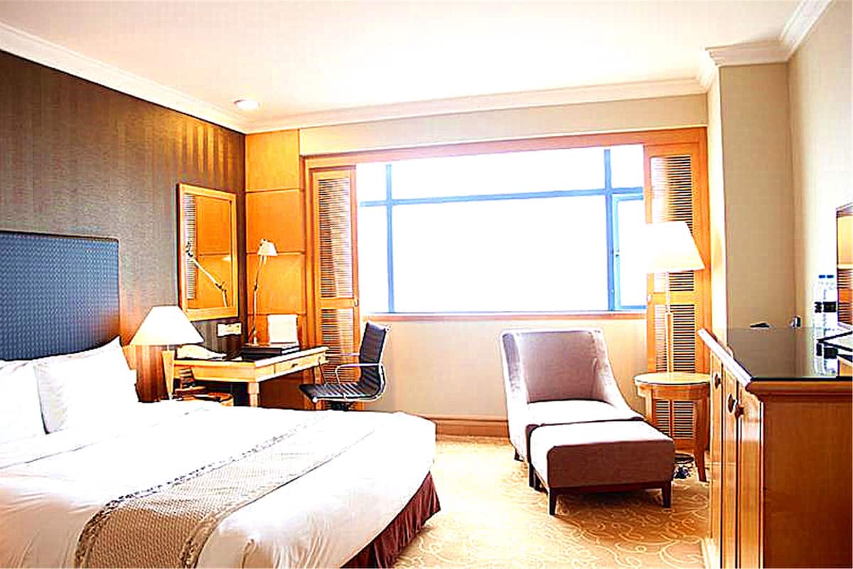 Habitación de hotel con una cama grande, un escritorio con una silla, una silla más pequeña y un sofá. Hay una ventana con persianas y un espejo en la pared.