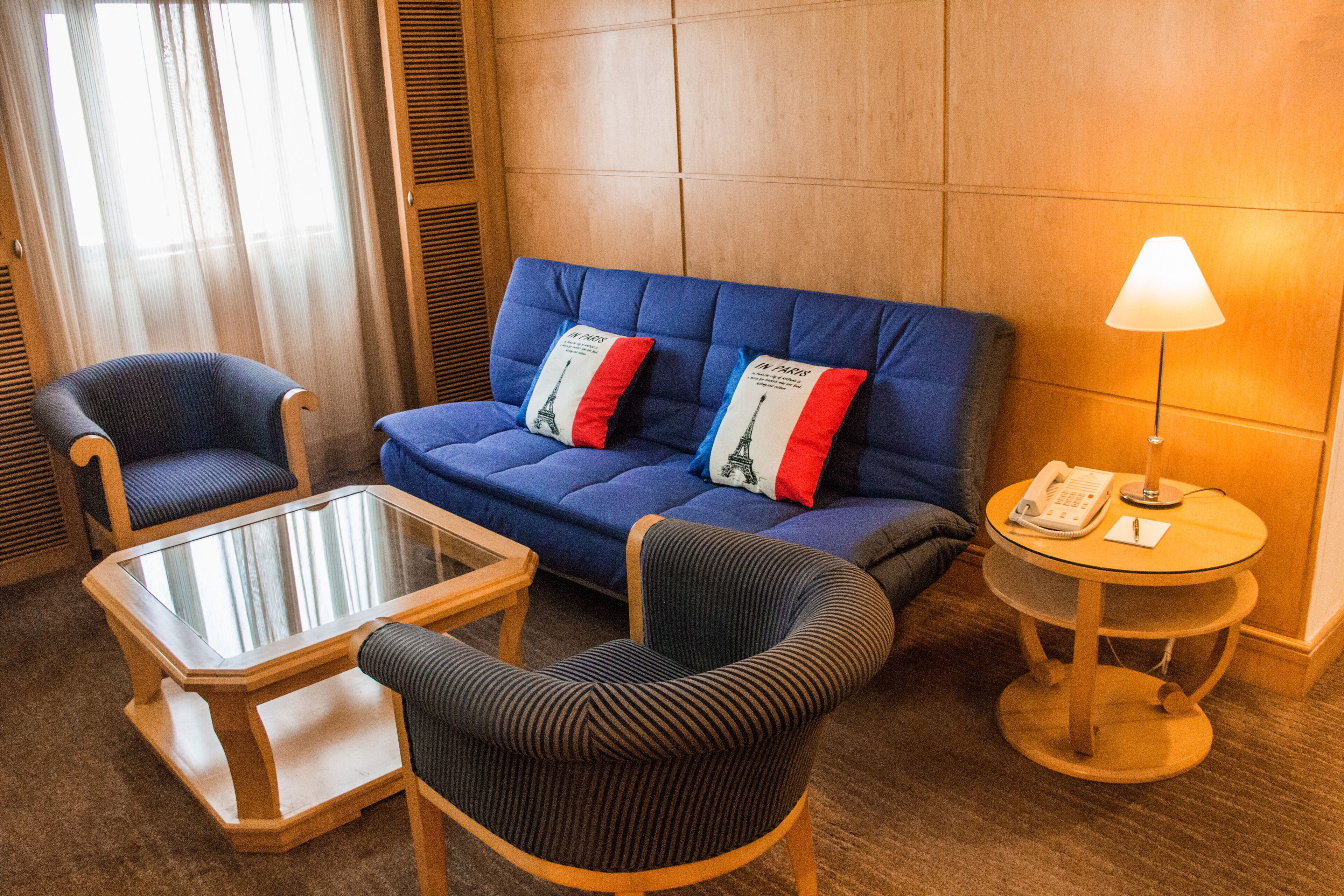 A hotel room with a blue sofa, two armchairs, a glass coffee table, a round side table with a lamp, and a telephone.