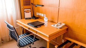 Habitación de hotel con un escritorio de madera, una silla de oficina negra, una lámpara de escritorio, un teléfono, un calendario y un cubo de basura.