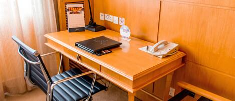 Habitación de hotel con un escritorio de madera, una silla de oficina negra, una lámpara de escritorio, un teléfono, un calendario y un cubo de basura.
