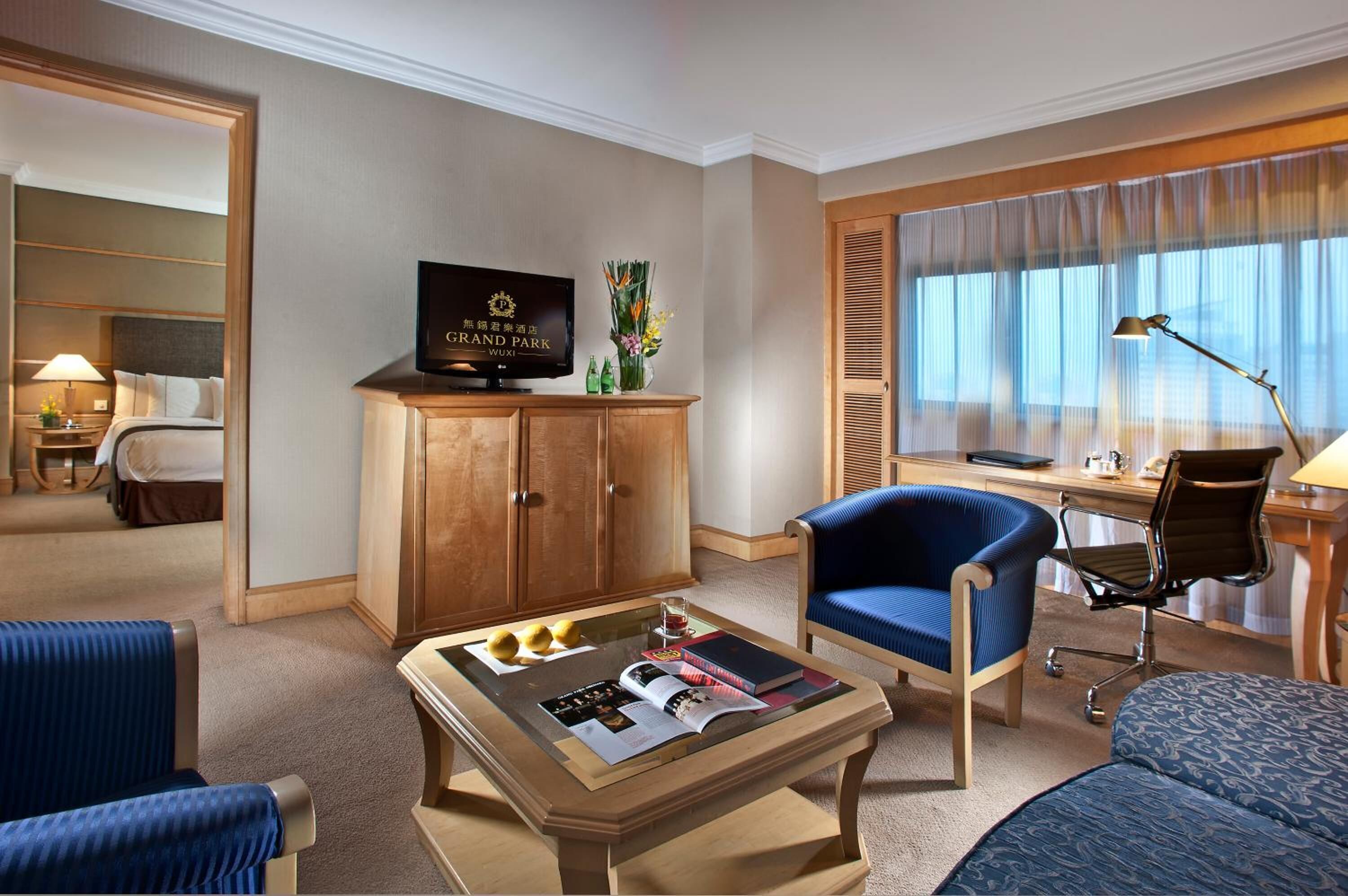 Habitación de hotel con cama, televisor en un mueble de madera, escritorio con silla, mesa de centro con revistas y una ventana grande con cortinas.