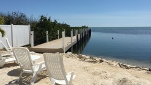 A beachside area with a wooden pier extending into calm waters, surrounded by white lounge chairs and a white fence.