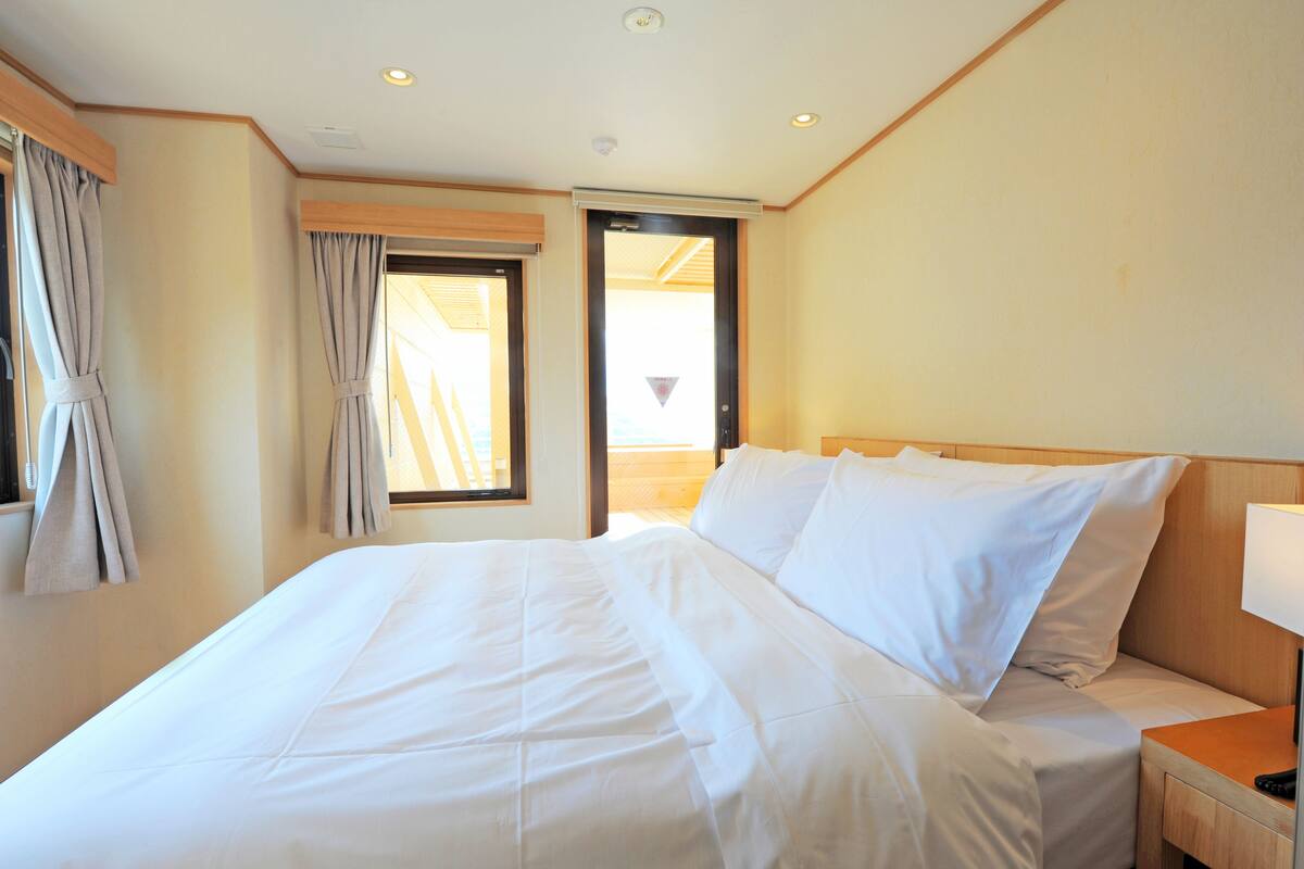 Una habitación de hotel con una cama grande, cabecera de madera, mesita de noche con lámpara y ventana con cortinas.