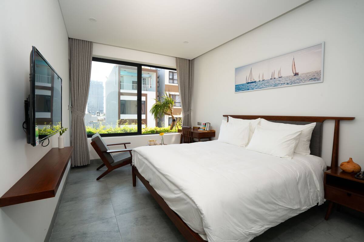 A modern hotel room with a large bed, a desk, a chair, a TV, and a window with a city view.