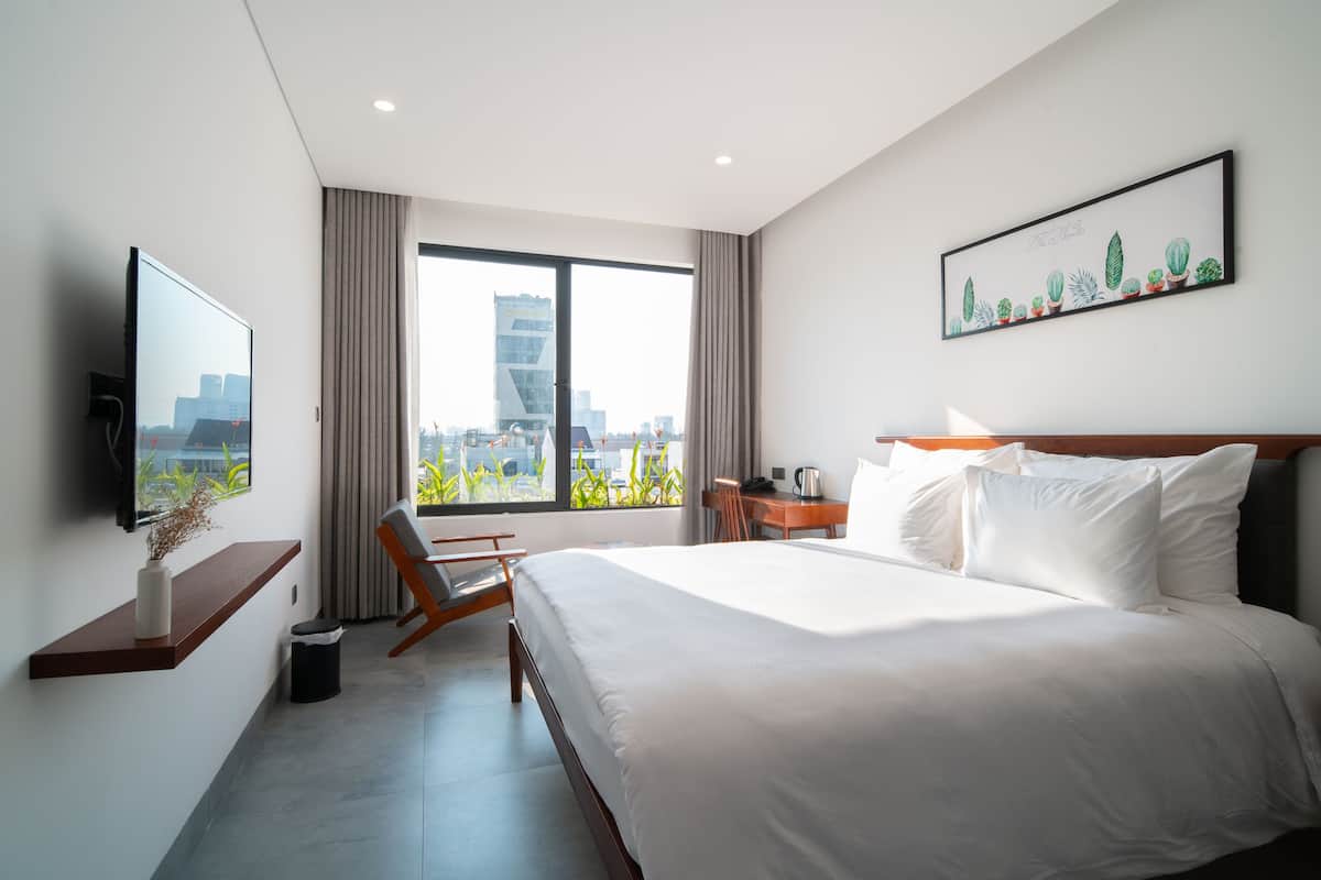 A modern hotel room with a large bed, a desk, a chair, a TV, and a window with a city view.