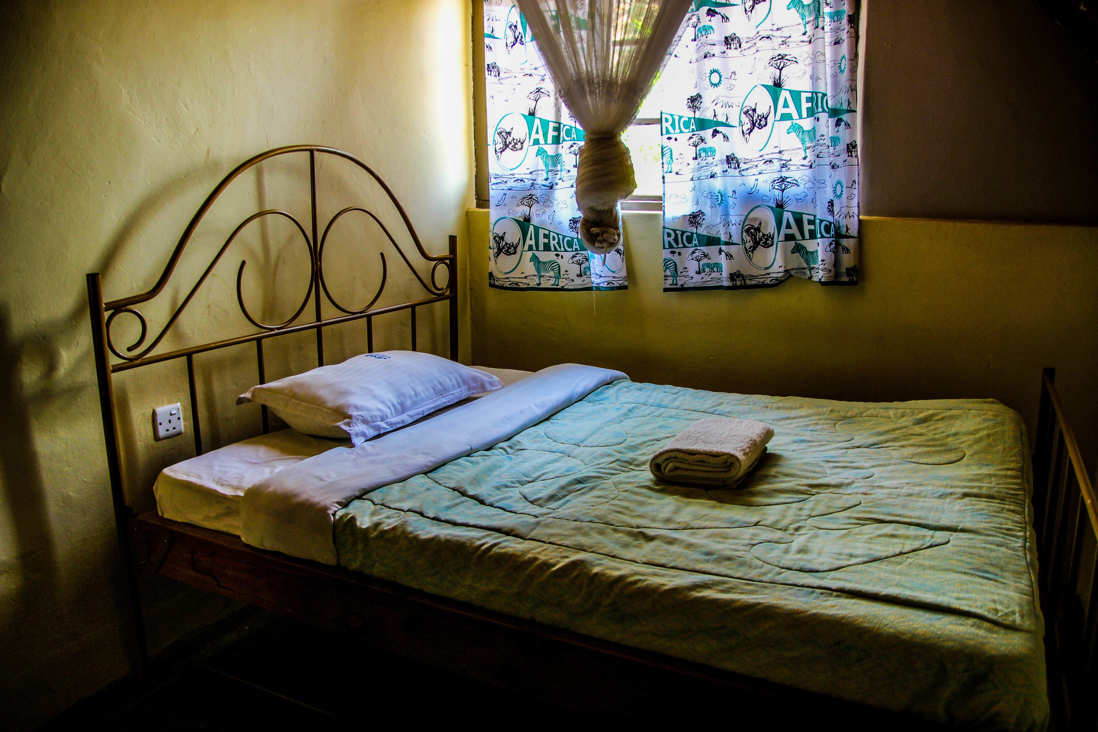Un lit simple avec une tête de lit en métal, une fenêtre ornée de rideaux à motifs et une table de chevet équipée d’une prise électrique.