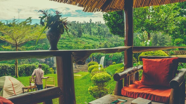 A wooden deck with a red cushioned chair and a table with books. A person is standing in the background. - Sipi Falls lodge (Sipi)
