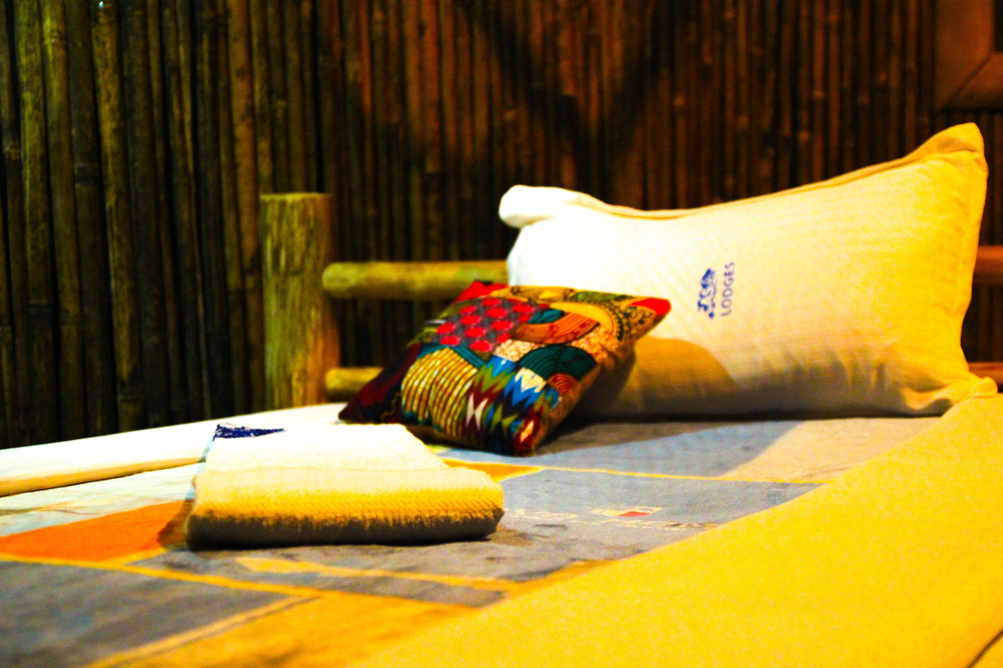 Una cama con un almohadón colorido y un almohadón liso, una manta enrollada y un cabecero de madera.