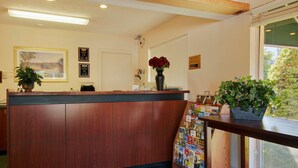A reception desk with a wooden finish, a vase with red flowers, a framed picture on the wall, and a window with a view of trees. - Knights Inn Salem (Salem)