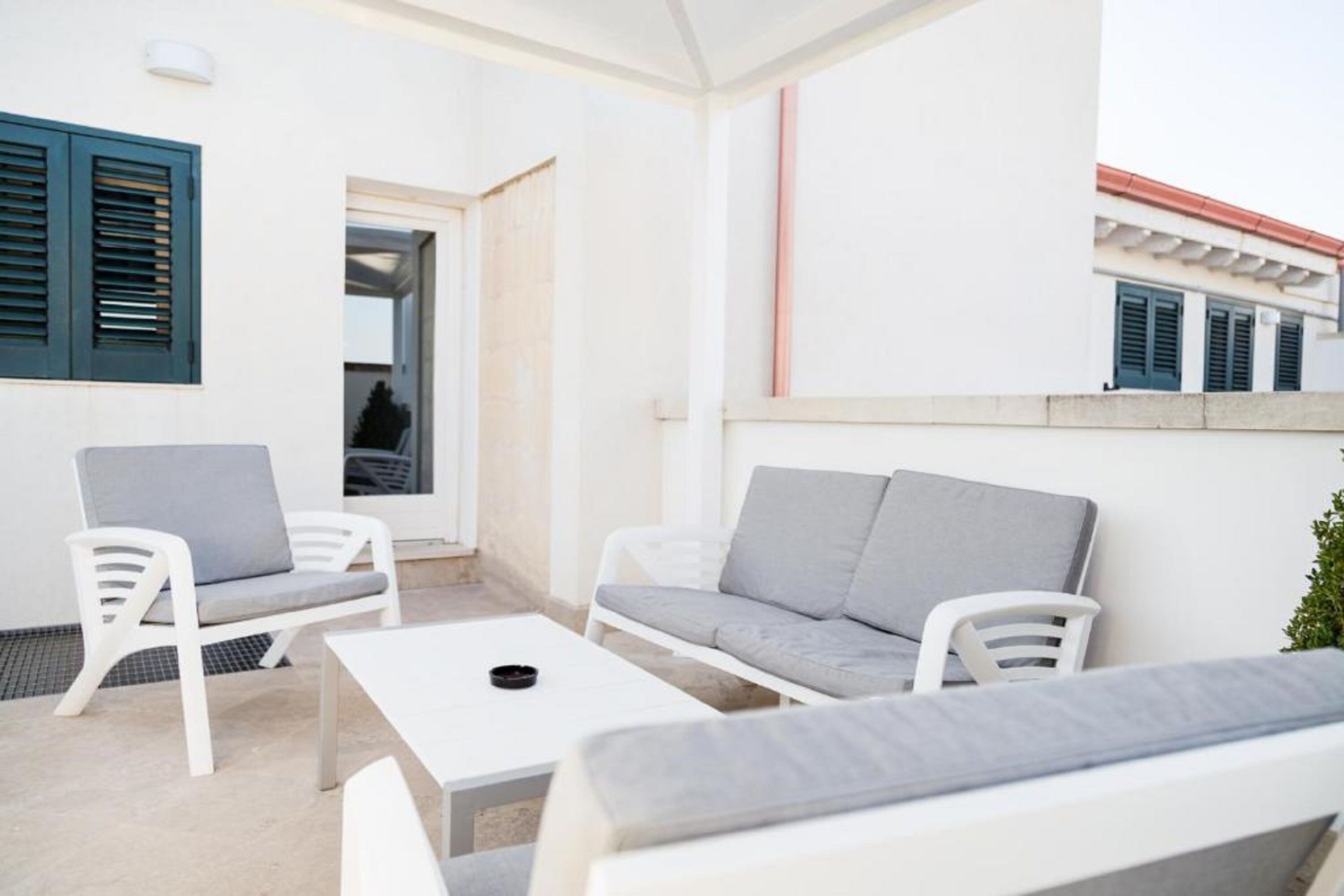 A modern outdoor seating area with white furniture and a small table, a door leading inside, and a building with a red roof in the background.