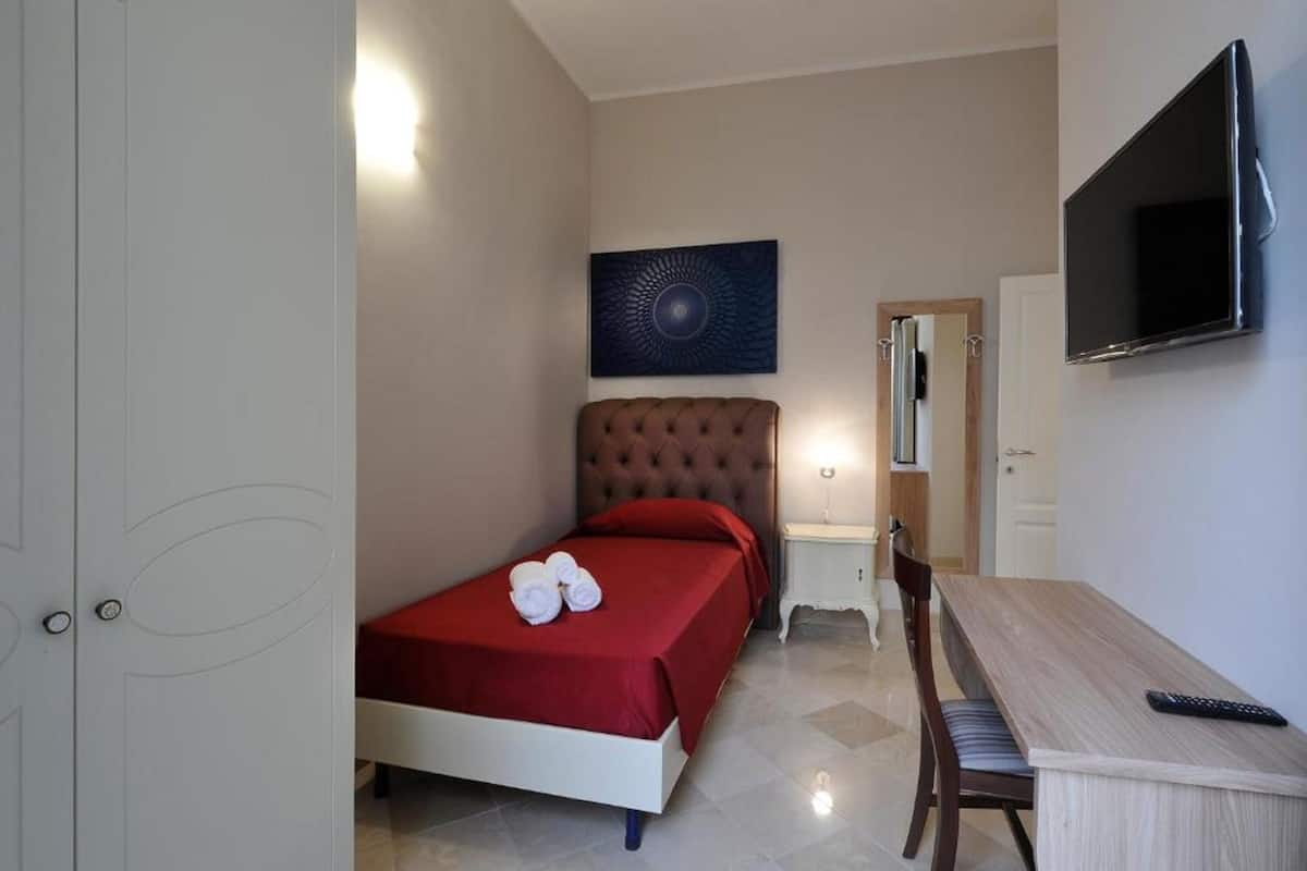 A bedroom with a bed, a desk, a chair, a TV, and a painting on the wall.