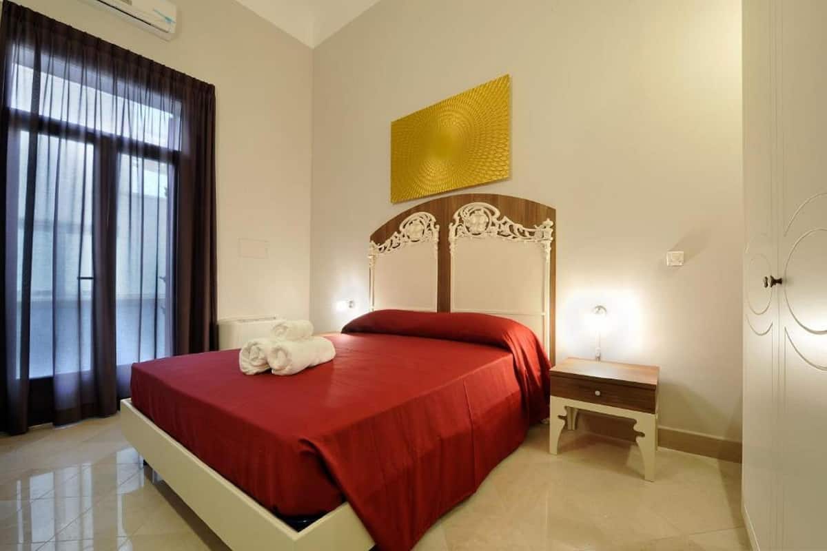 A hotel room with a bed, a nightstand, a wall-mounted artwork, and a window with curtains.