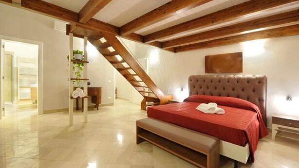 Un dormitorio con techo de madera, una cama con colcha roja, un banco, una escalera y un estante en la pared con plantas.
