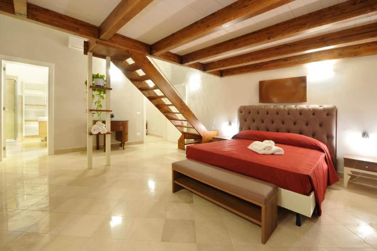 A bedroom with a wooden ceiling, a bed with a red bedspread, a bench, a staircase, and a wall-mounted shelf with plants.