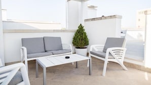 Una terraza en la azotea con un sofá blanco, dos sillas blancas, una mesita y una planta en maceta.