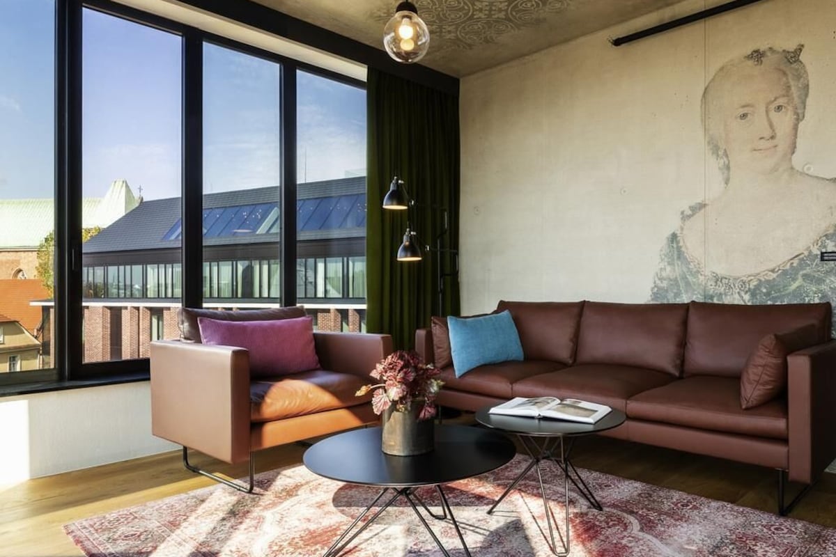 A modern living room with a large window, a brown sofa, a round coffee table, and a painting on the wall.