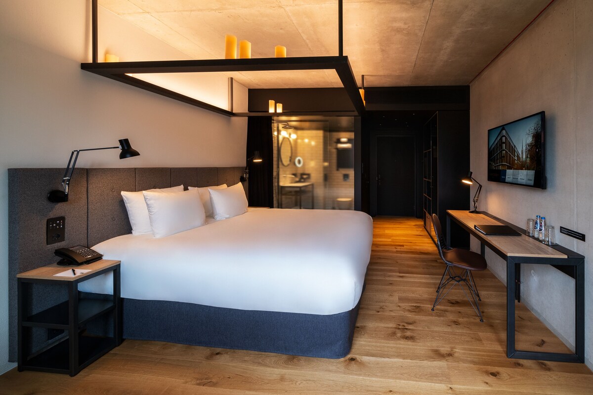 A modern hotel room with a large bed, bedside table, desk, and chair.