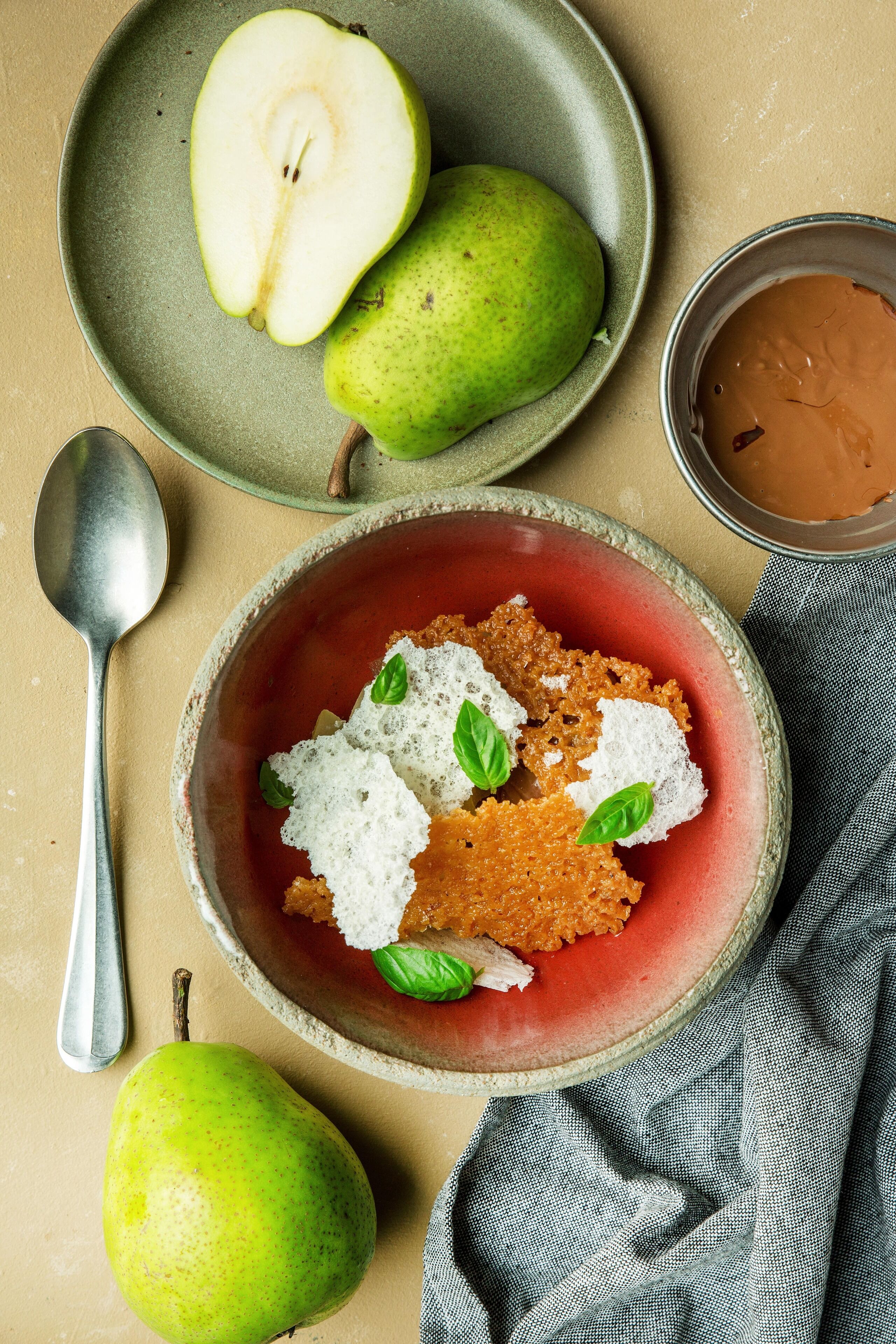 A bowl of food with a red base, garnished with white and green elements, accompanied by whole and halved pears on a plate.