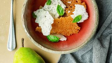 A bowl of food with a red base, garnished with white and green elements, accompanied by whole and halved pears on a plate.