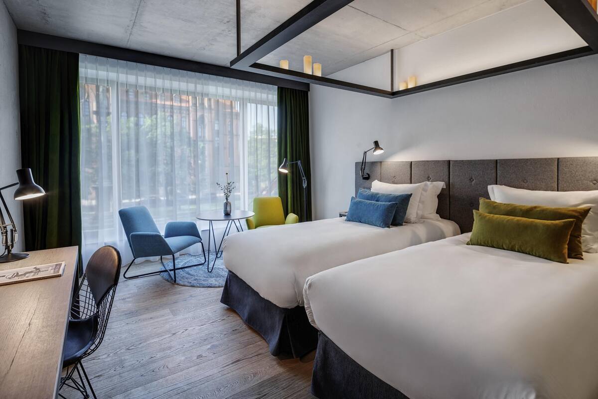 A hotel room with two beds, a desk with a chair, a small table with a vase, and a view of the outside through large windows.