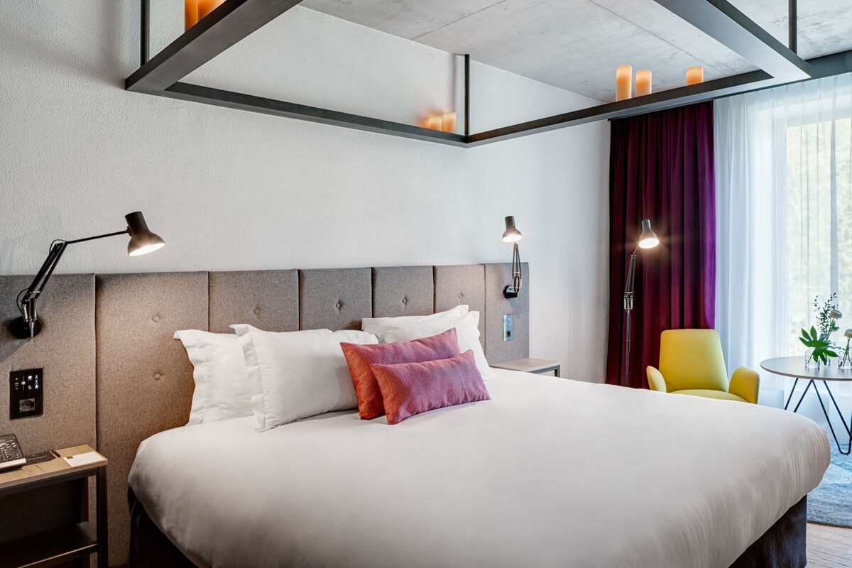 A modern hotel room with a large bed, bedside lamps, a small table with a vase, and a chair.