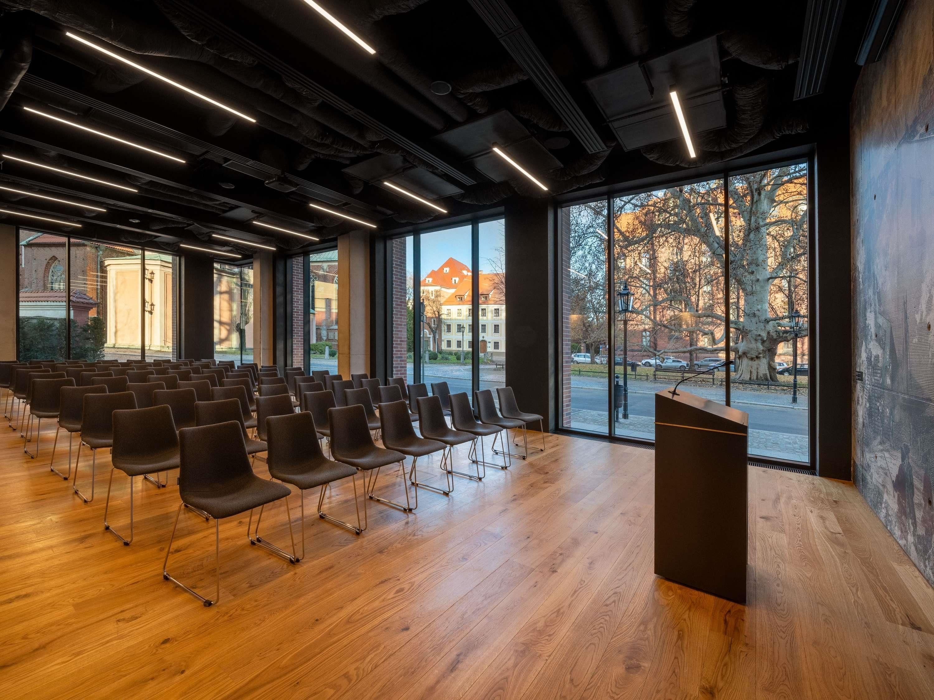 A conference room with rows of chairs, a podium, and large windows with a view of buildings and trees.