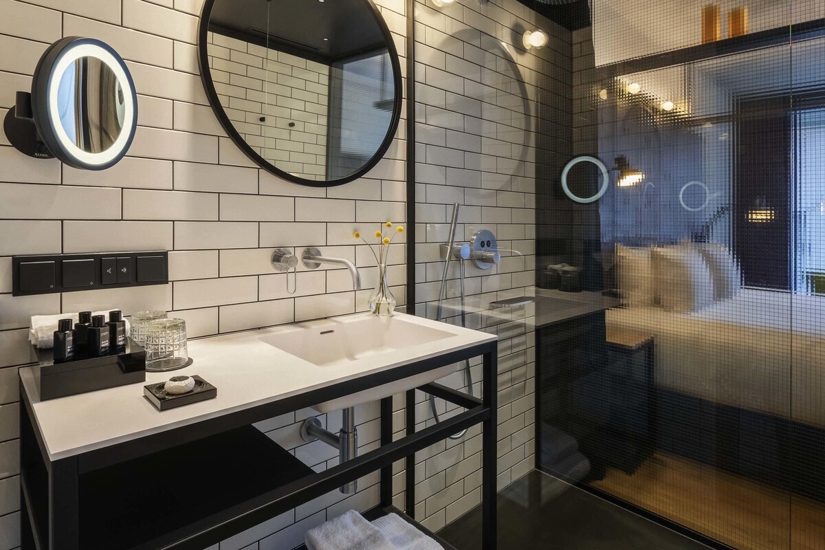 A modern bathroom with a white-tiled wall, a black vanity, a round mirror, and a glass-enclosed shower area.