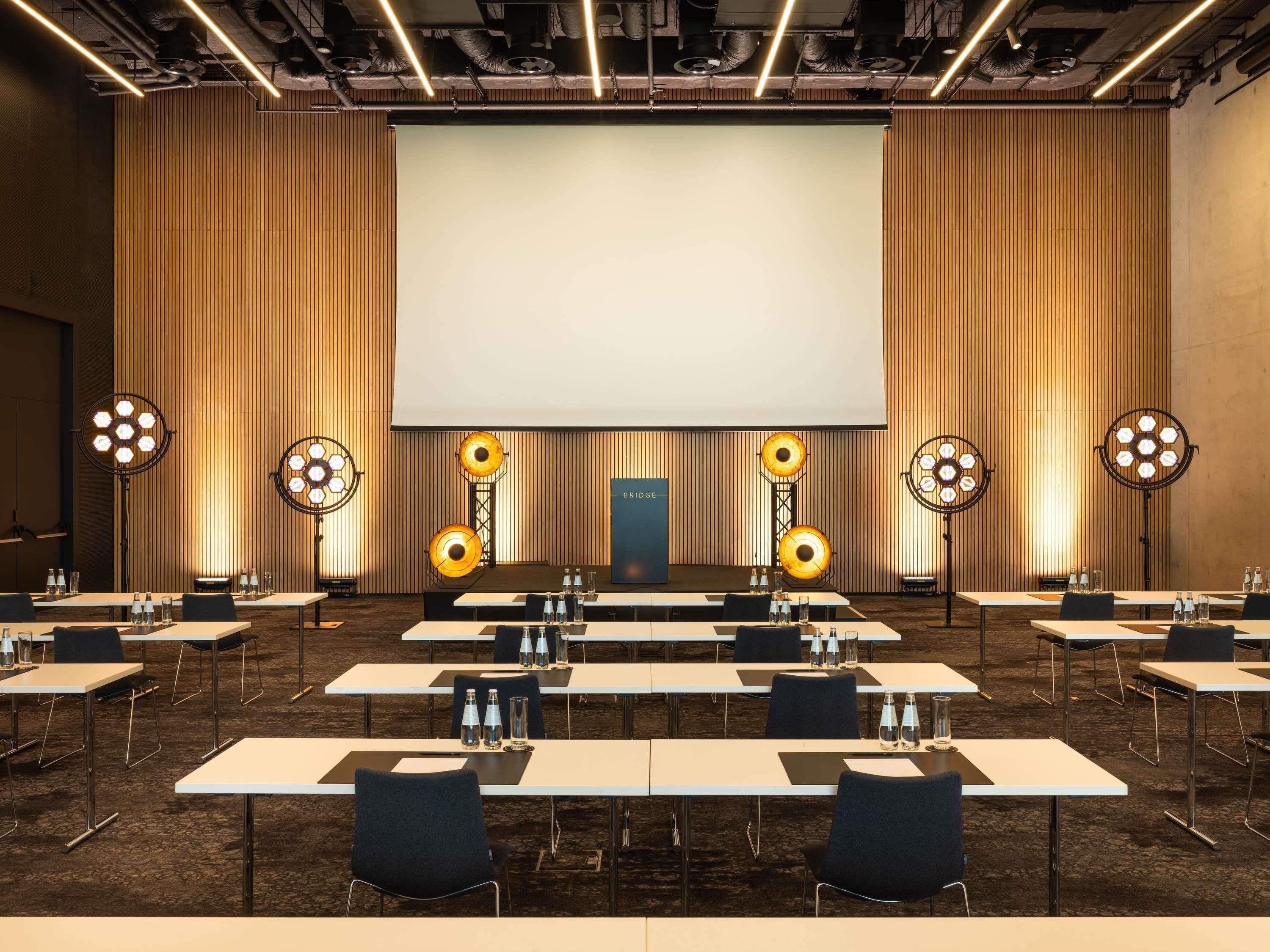 Une salle de conférence dotée d’un grand écran, de plusieurs tables, de chaises et d’éléments d’éclairage décoratifs.