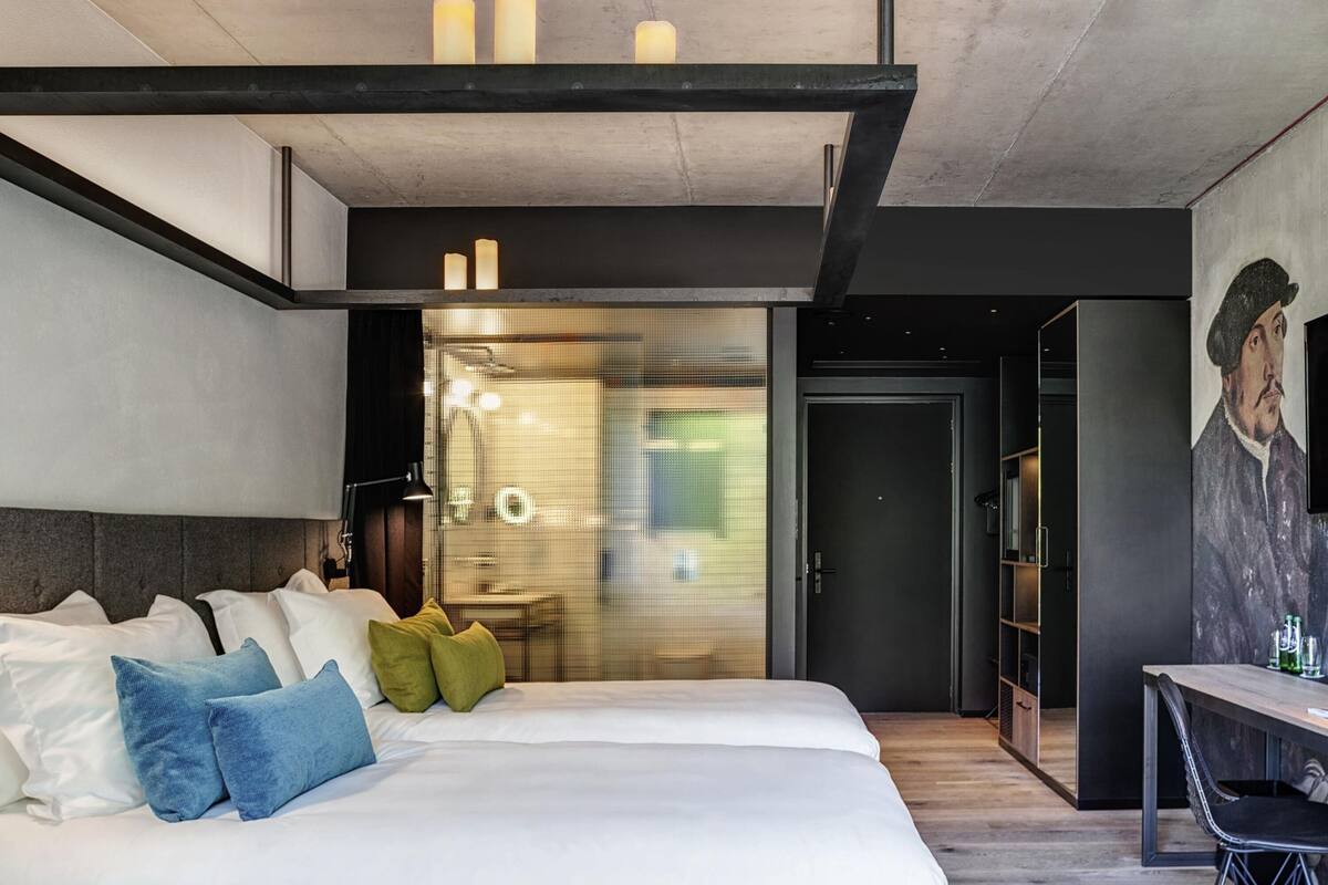 A modern hotel room with a large bed, a desk, and a painting on the wall.