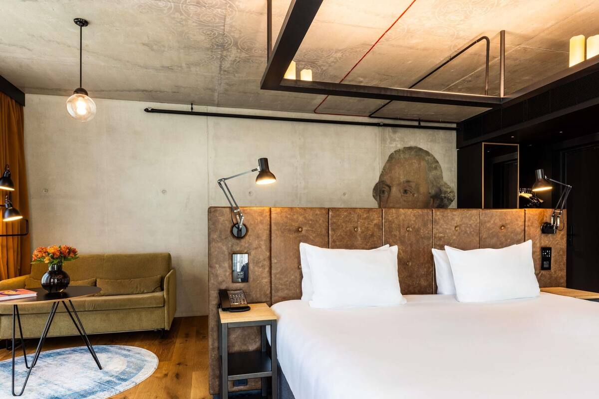 A modern hotel room with a large bed, a wooden headboard, a small side table with a lamp, a sofa, and a round rug.
