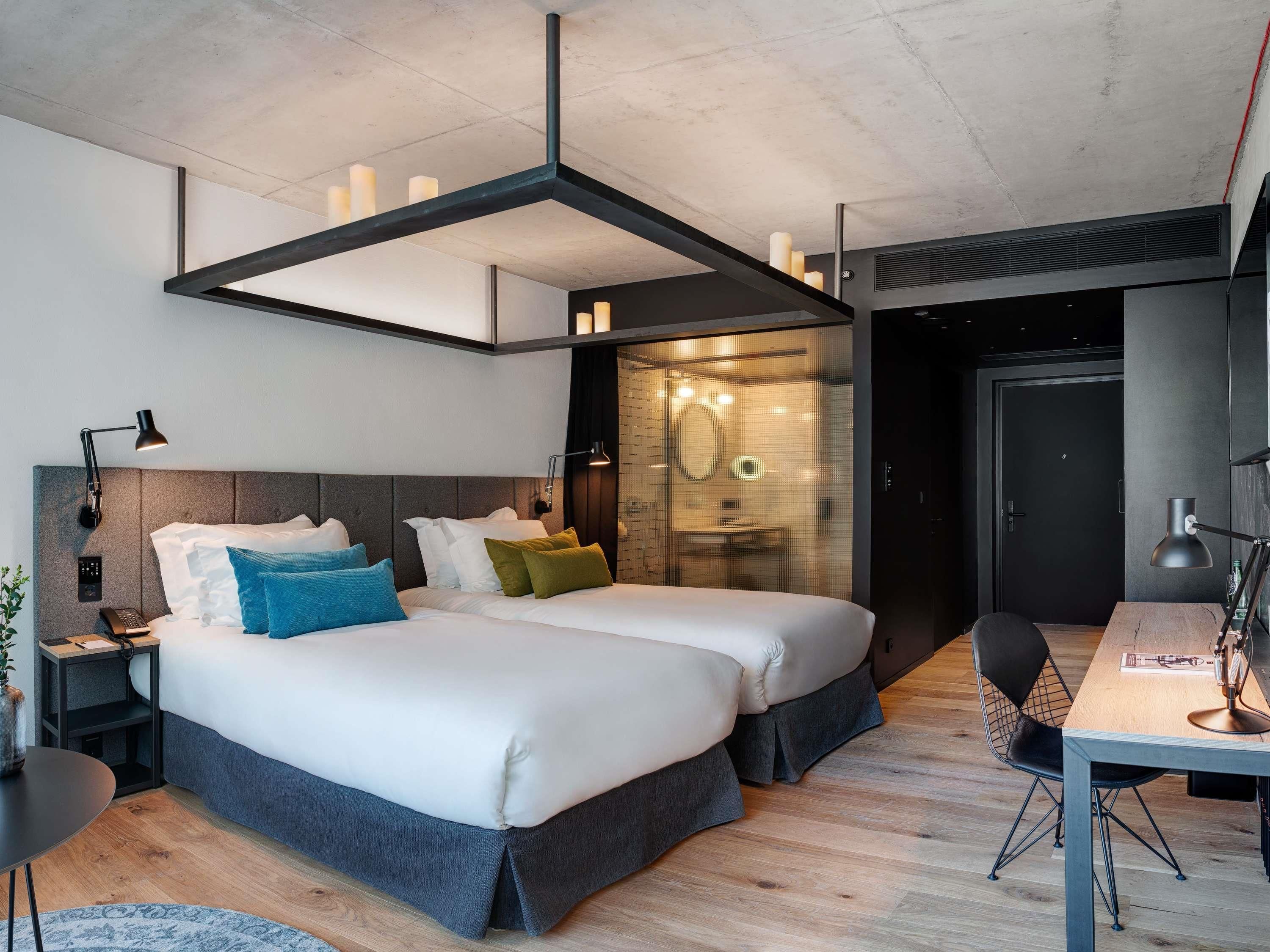 A modern hotel room with a large bed, bedside tables, a desk, and a bathroom visible through a glass partition.