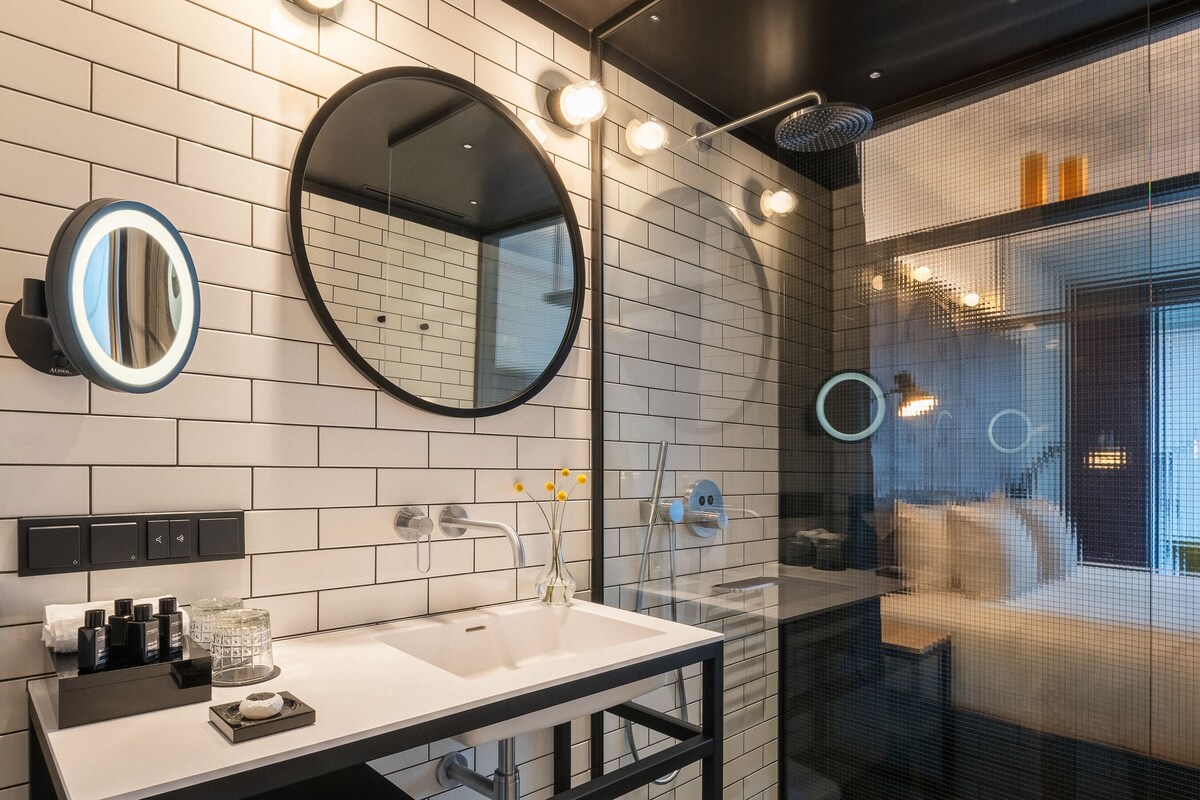 A modern bathroom with a white-tiled wall, a black vanity, a round mirror, and a glass-enclosed shower area.