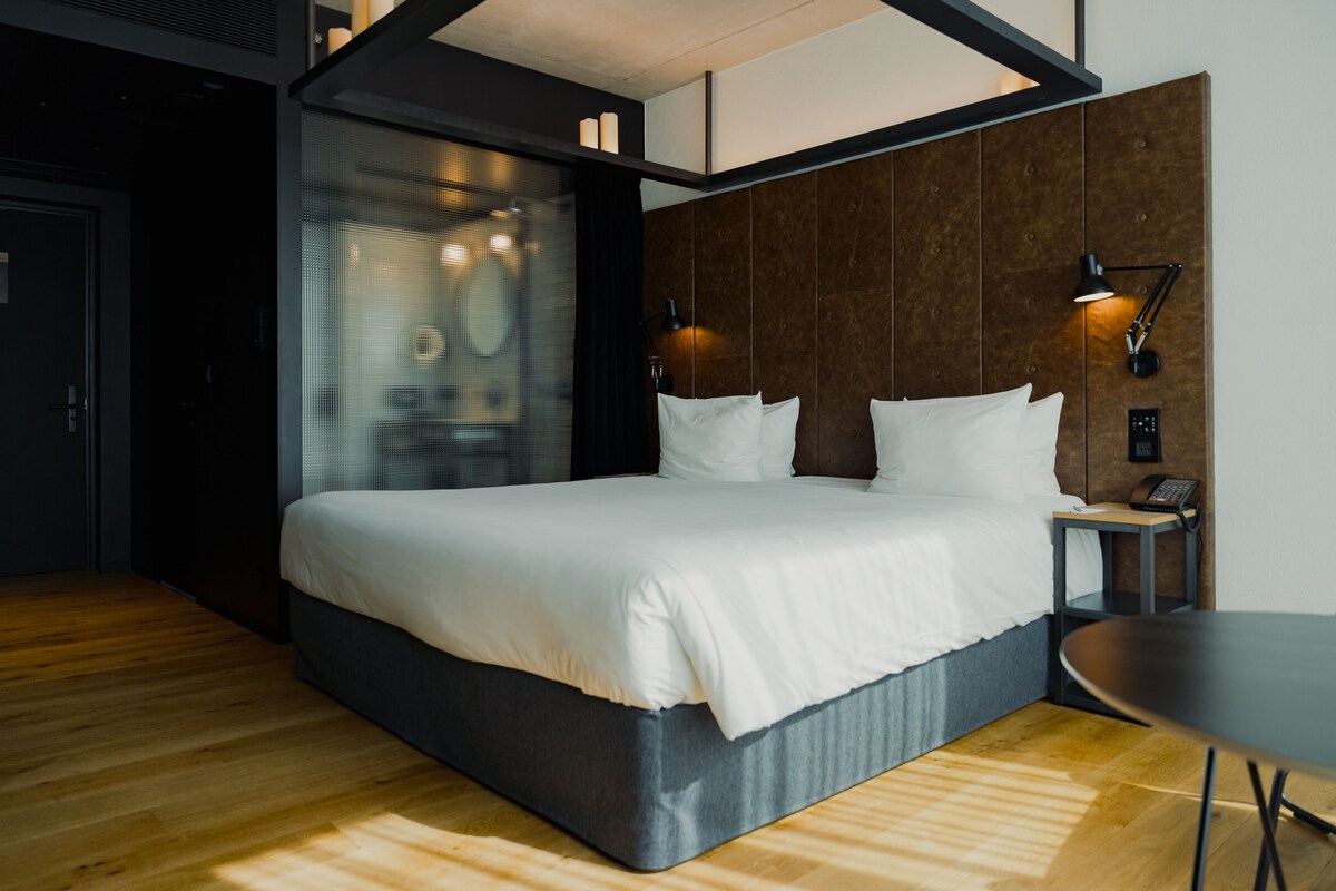 A modern hotel room with a large bed, a wooden floor, a wall-mounted TV, and a bedside table with a lamp.